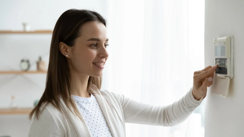 Femme souriante ajustant un thermostat numérique mural dans un intérieur moderne avec étagères flottantes en bois