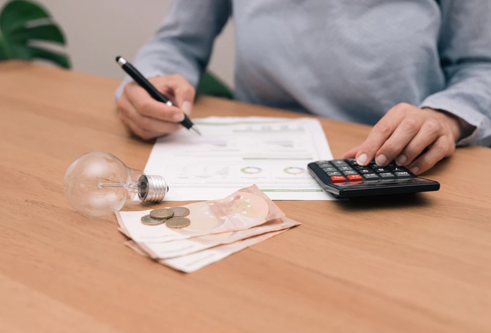 Gros plan sur une personne méconnaissable analysant une facture d'électricité avec une calculatrice, une ampoule et de l'argent sur un bureau